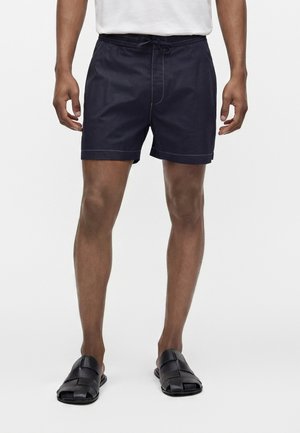 Homme portant un short bleu marine avec cordon, un t-shirt blanc et des sandales noires tissées ouvertes à l'avant, debout sur un fond clair uni.