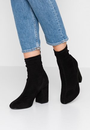 Personne portant des bottines en daim noir à talons blocs et un jean bleu court, debout sur un sol blanc lisse.