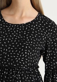 Vestido negro con escote redondo, mangas largas y cintura con volantes. La tela presenta un patrón de pequeños diseños en forma de hoja en color blanco.