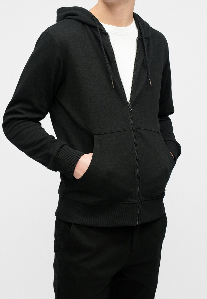 Personne portant un sweat à capuche noir zippé avec les mains dans les poches avant, superposé à un t-shirt blanc, debout devant un fond uni.