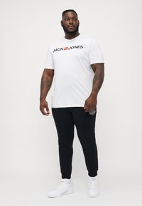 Witte katoenen t-shirt met zwart "JACK & JONES" logo, gecombineerd met zwarte joggers met een kleine graphic, en witte sneakers.