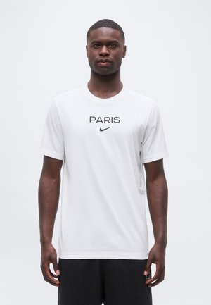 Fiatal férfi áll, fehér pólót visel "PARIS" felirattal és fekete Nike logóval a mellkasán, fekete rövidnadrággal párosítva.