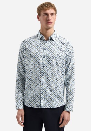 Jeune homme portant une chemise à manches longues à boutons avec un motif géométrique bleu, blanc et beige, associée à un pantalon foncé.