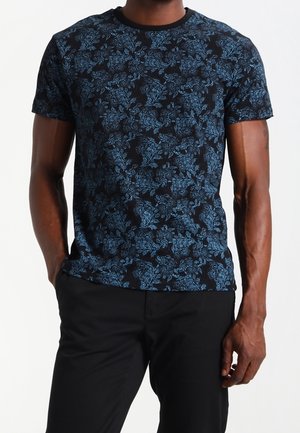 Homme portant un t-shirt noir à manches courtes avec un motif floral bleu sur toute la surface et un pantalon noir, debout les mains dans les poches.