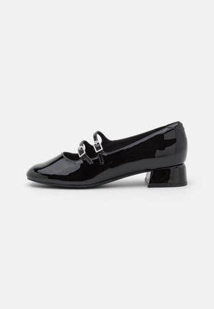 Chaussure à talon bas en cuir verni noir avec deux brides avant ajustables et boucles en argent, présentée en vue latérale sur un fond blanc.