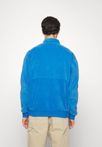 Felpa in pile blu con collo alto, polsini e vita a coste. Presenta una texture liscia e un dettaglio di cucitura orizzontale sul retro.