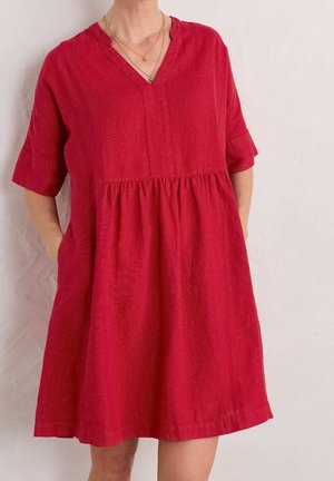 Nainen, joka pukee yllensä löysän, lyhythihaisen punaisen mekon, jossa on taskut ja V-kaula-aukko, ja asustaa kerroksellisilla kultakoruilla.