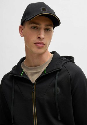 Felpa nera con zip, dettagli verde e un logo rotondo. Indossata sopra una maglietta beige, completata da un cappellino nero con una visiera cucita in oro.