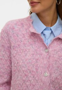 Rausvos spalvos megzta kardiganas su rombų raštu, šaliko apykakle ir sagų užsegimu, derinamas su šviesiai mėlyna marškinėlių apykakle po apačia.