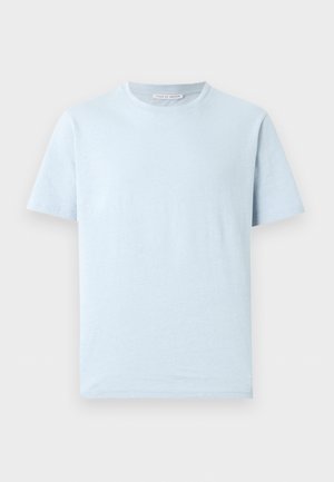 Homme debout avec les mains dans les poches, portant un t-shirt bleu clair, un pantalon bleu foncé à cordon et des baskets blanches sur un fond neutre.