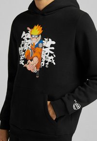 Sudadera negra de tejido suave con un diseño gráfico colorido de Naruto en la parte delantera y un bolsillo tipo canguro. Detalle blanco en la manga.