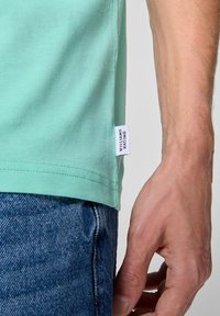 Jasnozielona bawełniana koszulka z krótkim rękawem, z rozcięciem po bokach, z białą metką z napisem "Williams Racing" widoczną na dole. Nosi się ją z ciemnymi dżinsami.