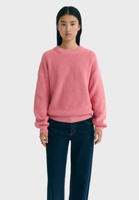 Pull en tricot rose avec une texture côtelée, col rond et épaules tombantes, associé à un jean foncé, sur un fond uni.