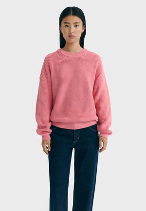 Roze gebreide trui met een geribde textuur, ronde halslijn en verlaagde schouders, gecombineerd met donkere jeans, op een effen achtergrond.