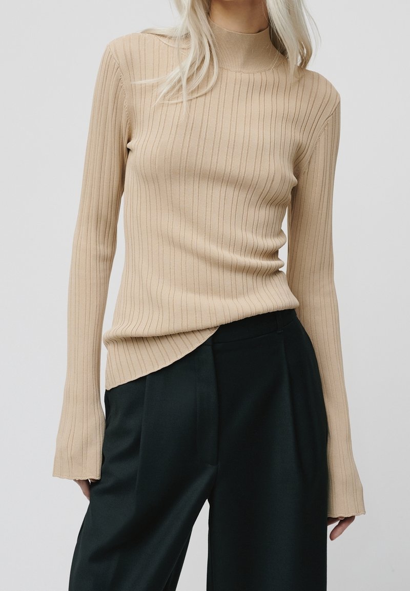 Haut beige côtelé à manches longues avec un col montant et des manches allongées, associé à un pantalon large vert foncé.