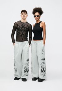 Mustast pika varrukaga võrgust pluus lillekujundusega, paaritatud hallide püksidega, millel liblikate graafika ja tekst. Mõlemad kannavad musti jalatseid.