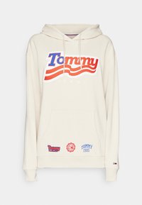 Sweat à capuche crème avec un grand logo "Tommy" en rouge et bleu, poche kangourou à l'avant avec trois petites étiquettes de marque Tommy Jeans, et capuche avec cordon de serrage.