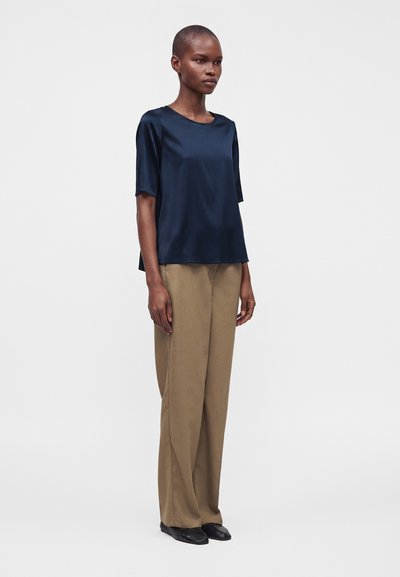 Femme debout portant un chemisier en satin bleu foncé à manches courtes, un pantalon beige ample et des chaussures noires sur un fond clair uni.