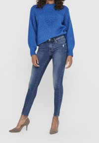 Suéter azul de punto con patrón de hojas, combinado con jeans ajustados azul deslavado y zapatos de tacón altos gris claro, posando contra un fondo blanco.