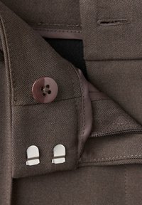 Gros plan d’un tissu brun avec un bouton brun foncé, deux crochets argentés pour la fixation, et des coutures visibles près d’une fermeture éclair.