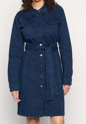 Femme portant une robe en jean bleu foncé longueur genou avec manches longues, fermeture boutonnée à l'avant, poches poitrine et ceinture assortie en tissu.