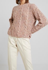Femme portant un pull en grosse maille torsadée rose et beige et un pantalon taille haute crème, debout les mains sur les hanches devant un fond uni.