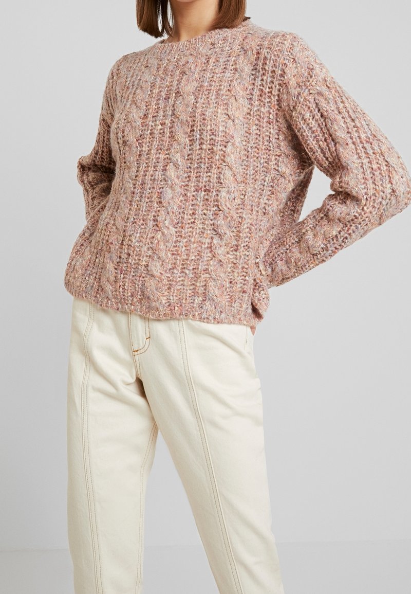 Femme portant un pull en grosse maille torsadée rose et beige et un pantalon taille haute crème, debout les mains sur les hanches devant un fond uni.