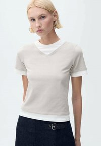 Tricou bej cu decolteu în V din material moale, cu mâneci scurte cu manșete albe, purtat peste un top alb cu guler rotund.
