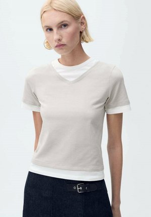 Beige V-neck t-shirt in soft fabric, featuring short sleeves with white cuffs, layered over a white crew neck top.