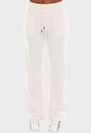 Pantalon de survêtement blanc ample avec taille à cordon porté par une personne debout, associé à des baskets blanches et grises.