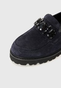 Mørkeblå semsket loafers med rund tå, gummisåle og tre sorte juvelakenter på overdelen. Har sydd detaljer langs kantene.