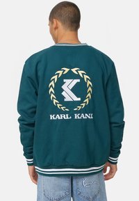 Felpa verde con logo ricamato in bianco e oro, con una corona di alloro. I polsini e l'orlo striati aggiungono un contrasto in blu e bianco.
