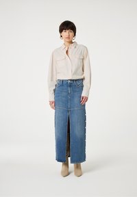 Denim-Maxikleid mit seitlichen Knöpfen, tiefer vorderer Schlitze und hoher Taille, kombiniert mit einer leichten, knopfbaren Bluse. Lederstiefeletten runden den Look ab.