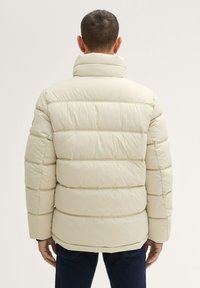 Chaqueta de plumas beige con secciones acolchadas horizontales, cuello alto y textura suave. La vista trasera muestra acentos acanalados a lo largo de los bordes.