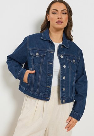 Vrouw draagt een donkerblauwe spijkerjack over een wit shirt en beige broek met een hoge taille, met één hand in de jaszak.