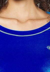Blå sammetsoberdel med rund halsringning och en metallkedjeaccent. Slät textur med lätt glans och en broderad logotyp på sidan.