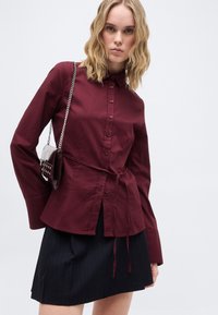Νέα γυναίκα με κυματιστά ξανθά μαλλιά φοράει πουκάμισο βουργουνδί με κουμπιά και ζώνη στη μέση, μαύρη φούστα και κρατά μικρή σκοτεινή τσάντα με αλυσίδα.
