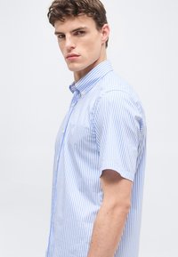 Νέος άνδρας που φοράει πουκάμισο με κοντά μανίκια σε ανοιχτό μπλε και λευκές κάθετες ρίγες, κοιτάζει πάνω από τον ώμο του με φόντο ένα απλό φόντο.