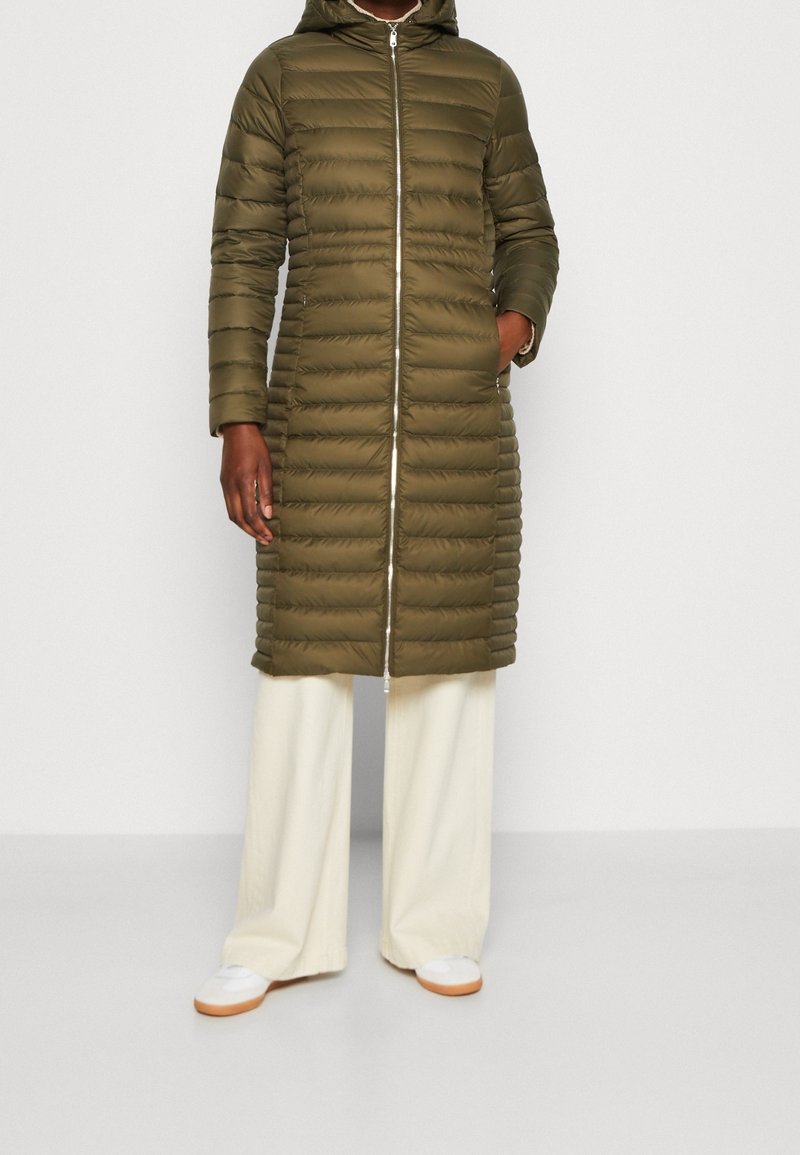 Persoon in een olijfgroene lange pufferjas met een capuchon, crème wijde broek en witte sneakers met rubberen zolen. Een hand in de zak.