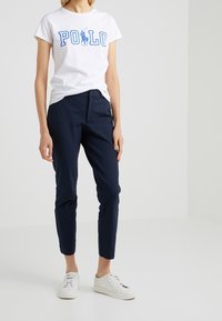 Vit t-shirt med blå "POLO"-tryck, marinblå slim-fit byxor och vita sneakers. T-shirten har korta ärmar; byxorna har fickor och rak passform.