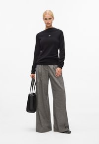 Schwarzer Pullover mit einem Bootsausschnitt und einem kleinen Logo. Graue, weit geschnittene Hose aus strukturiertem Stoff. Das Model hält eine schwarze Tasche.