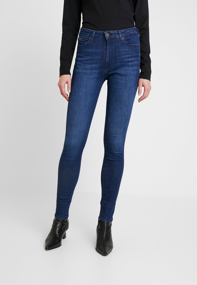 Mujer vistiendo jeans ajustados azul oscuro, top negro de mangas largas y botas de tacón negro con punta en un fondo liso.