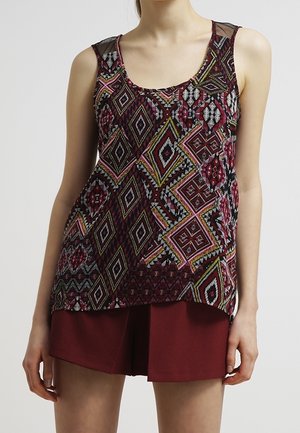 Donna con top senza maniche con motivi geometrici colorati e pantaloncini rosso scuro, in piedi contro uno sfondo chiaro e uniforme.