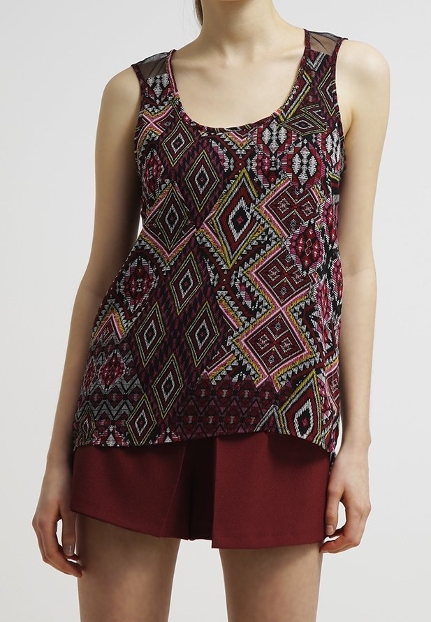 Femme portant un haut sans manches avec des motifs géométriques colorés et un short rouge foncé, debout devant un fond uni clair.