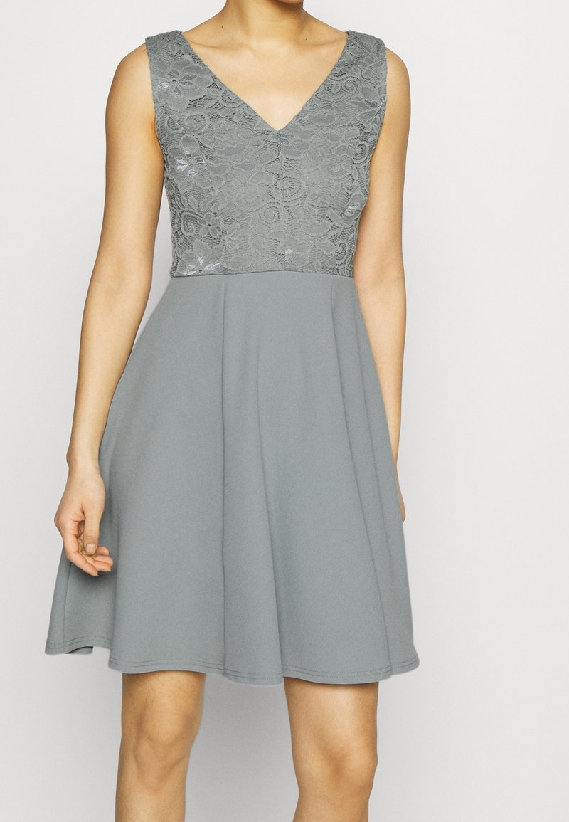 Robe grise sans manches avec un corsage en dentelle à col en V et une jupe évasée uni, portée par une personne debout.