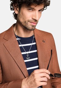 Brązowa blazer z teksturą nałożona na granatową koszulę w paski, z srebrnym wisiorkiem w kształcie kotwicy na łańcuszku. Trzyma czarne okulary przeciwsłoneczne.