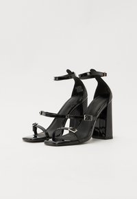 Sandales à talons hauts en cuir verni noir avec une bride de cheville, un bout carré et un talon épais. Présente des accents à boucle réglable.