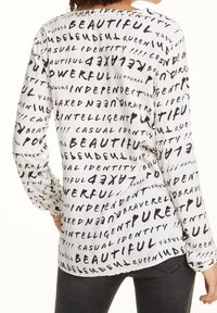 Langärmlige Bluse aus weißem Stoff mit einem schwarzen abstrakten Textmuster, das Wörter wie "schön" und "unabhängig" zeigt. Lockere Passform.