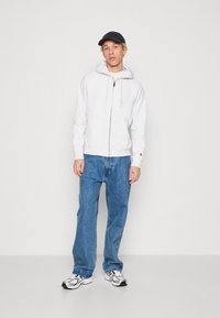 Hellgrauer Reißverschluss-Hoodie mit Kordeln, getragen über einem weißen Shirt. Kombiniert mit lockeren blauen Jeans und grauen Sneakers. Die Mütze ist marineblau.