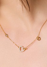 Złoty naszyjnik z łańcuszka z centralnym sercowym wisiorkiem wysadzanym przezroczystymi kamieniami oraz mniejszym serduszkowym urokiem. Gładka faktura.